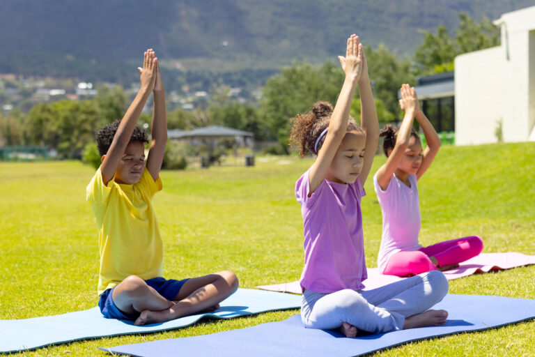 Biracial children practice yoga outdoors, sitting on colorful mats. The 9-year-old girl in purple leads the 8-year-old boy in yellow and the 10-year-old girl in pink in a peaceful pose.