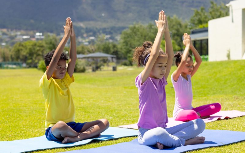 Biracial children practice yoga outdoors, sitting on colorful mats. The 9-year-old girl in purple leads the 8-year-old boy in yellow and the 10-year-old girl in pink in a peaceful pose.